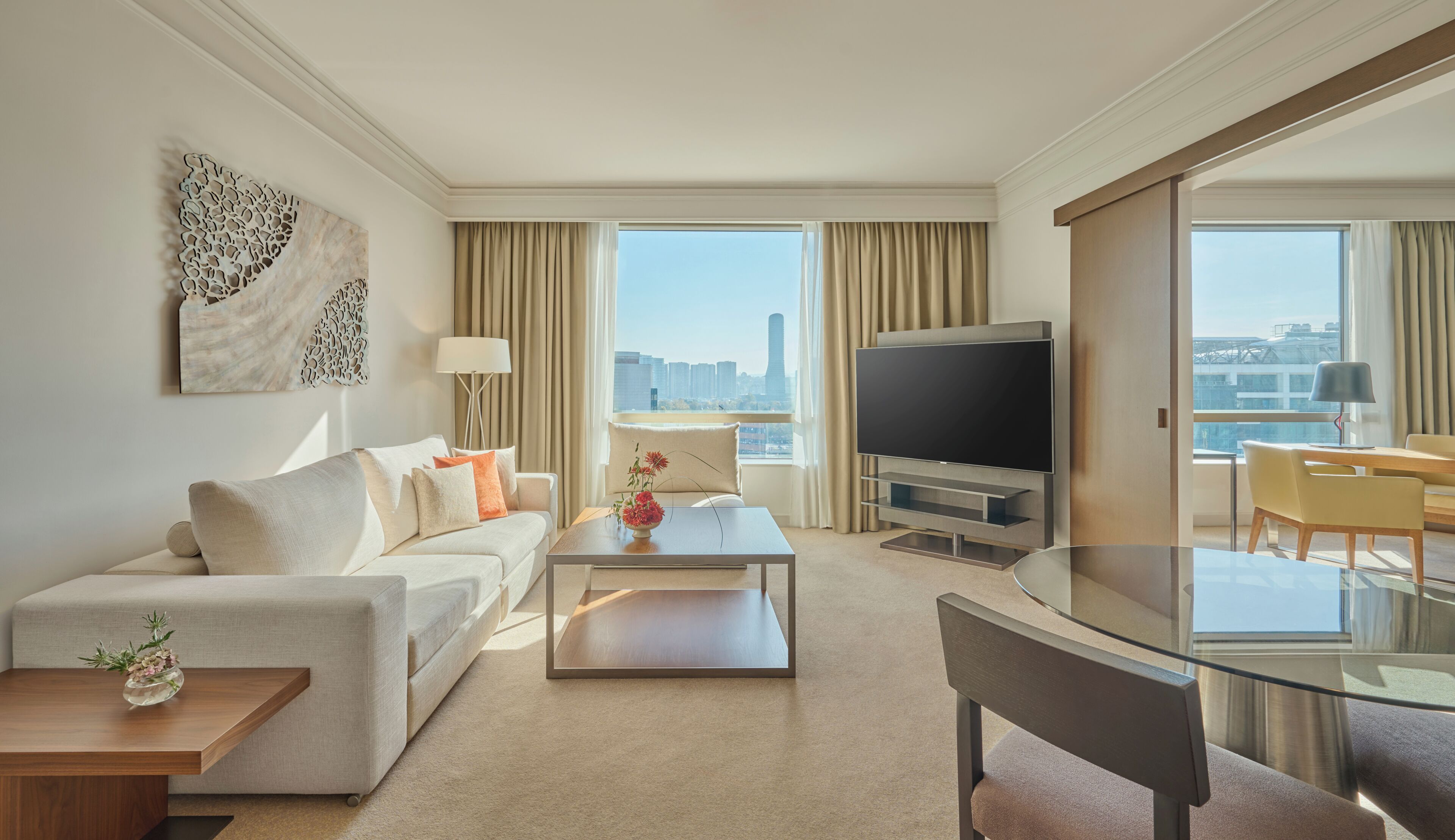 Quarto de hotel moderno com sofá, mesa de centro, televisão e área de refeições com mesa de vidro.