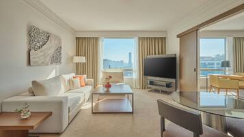 Quarto de hotel moderno com sofá, mesa de centro, televisão e área de refeições com mesa de vidro.