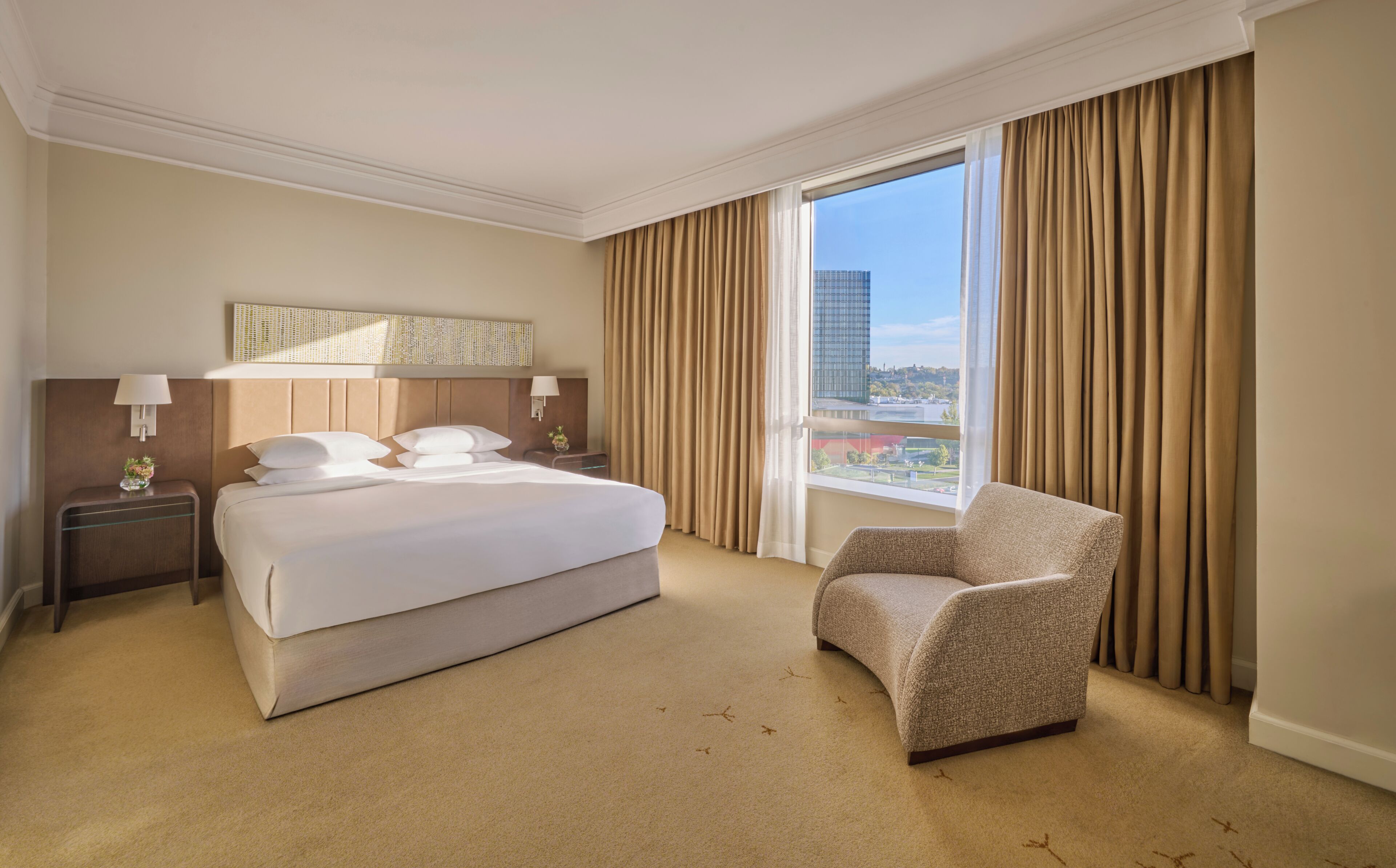 Habitación de hotel con una cama grande, una silla, una mesita de noche, una lámpara y vistas a un paisaje urbano a través de un ventanal amplio.