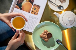 A person holding a cup of tea, a teapot, a plate with a decorative item, and a spoon on a table with a magazine.