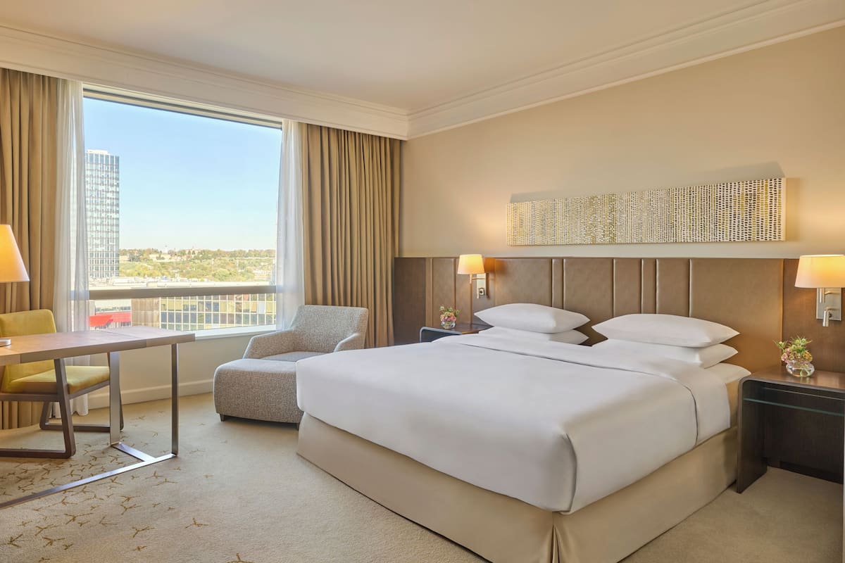 A hotel room with a large bed, a desk, a chair, a nightstand, and a view of the cityscape.