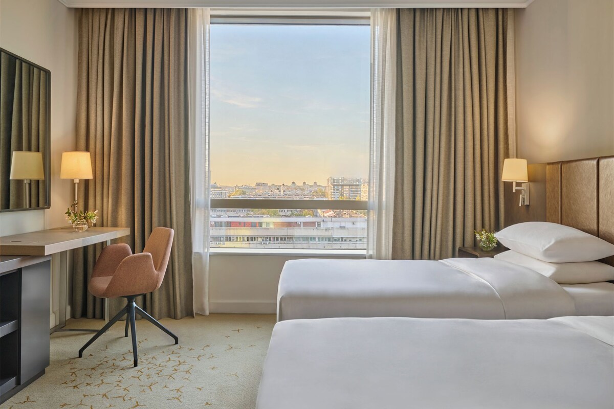 A hotel room with two beds, a desk, a chair, and a large window with a city view.