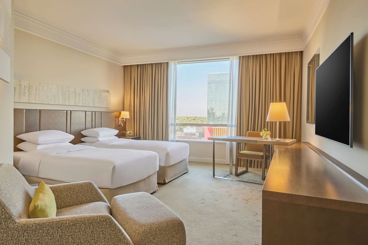 A hotel room with two beds, a desk, a TV, and a view of the city.