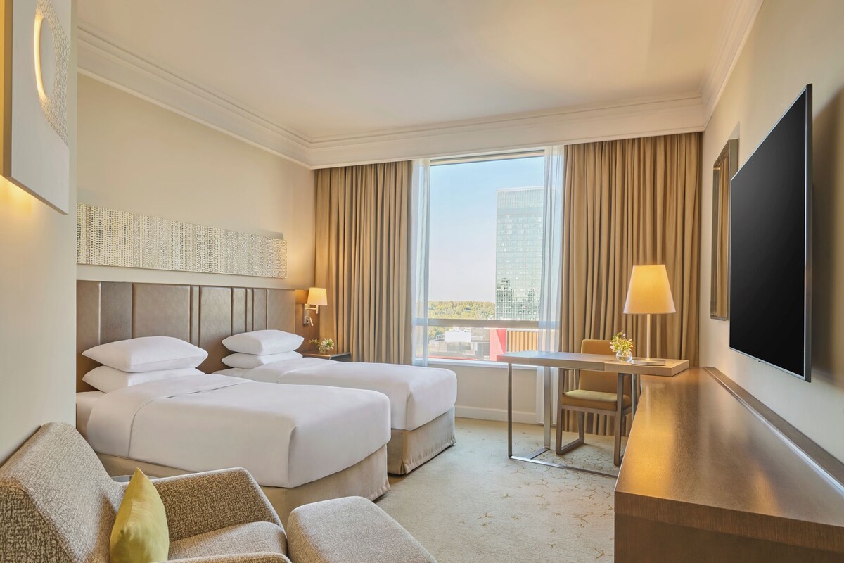 A hotel room with two beds, a desk, a chair, a TV, and a balcony with curtains.