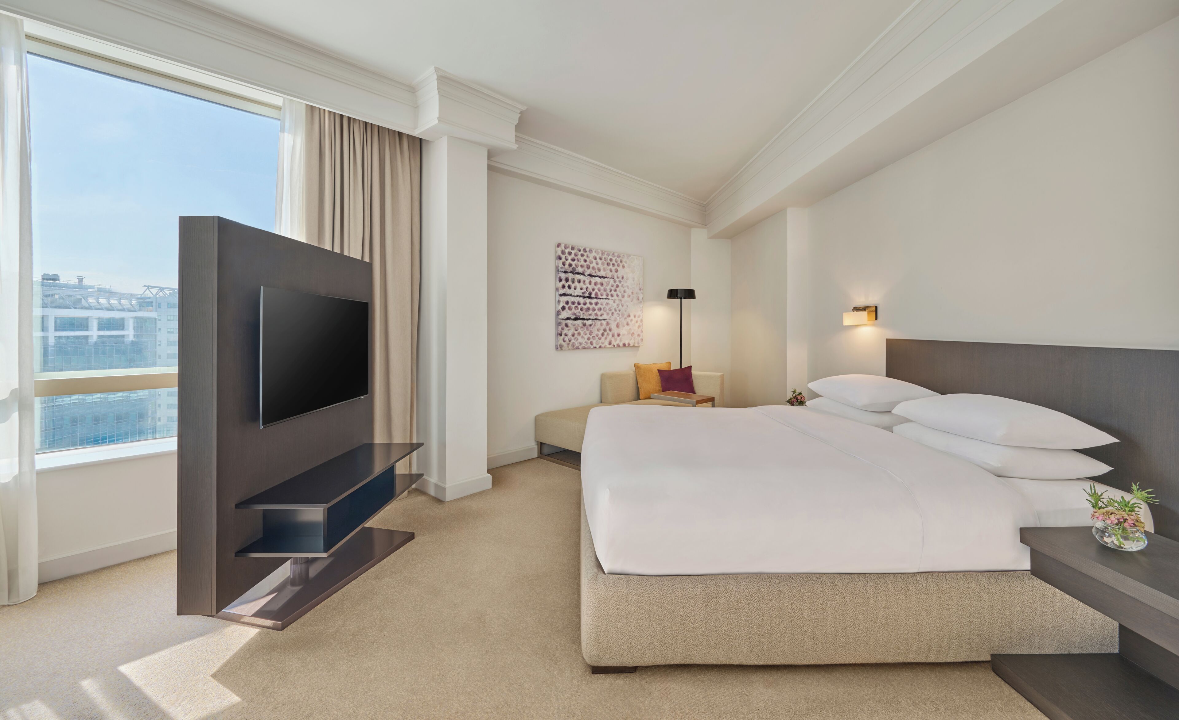 A hotel room with a bed, a TV mounted on a wall, a window with curtains, a wall-mounted light, and a view of buildings outside.