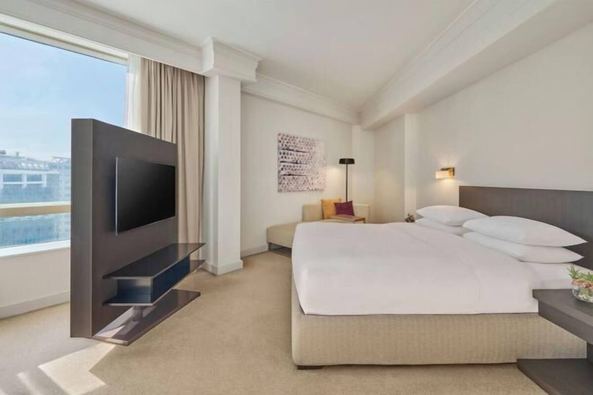 A hotel room with a bed, a TV mounted on a wall, a bedside table, a lamp, and a window with curtains.