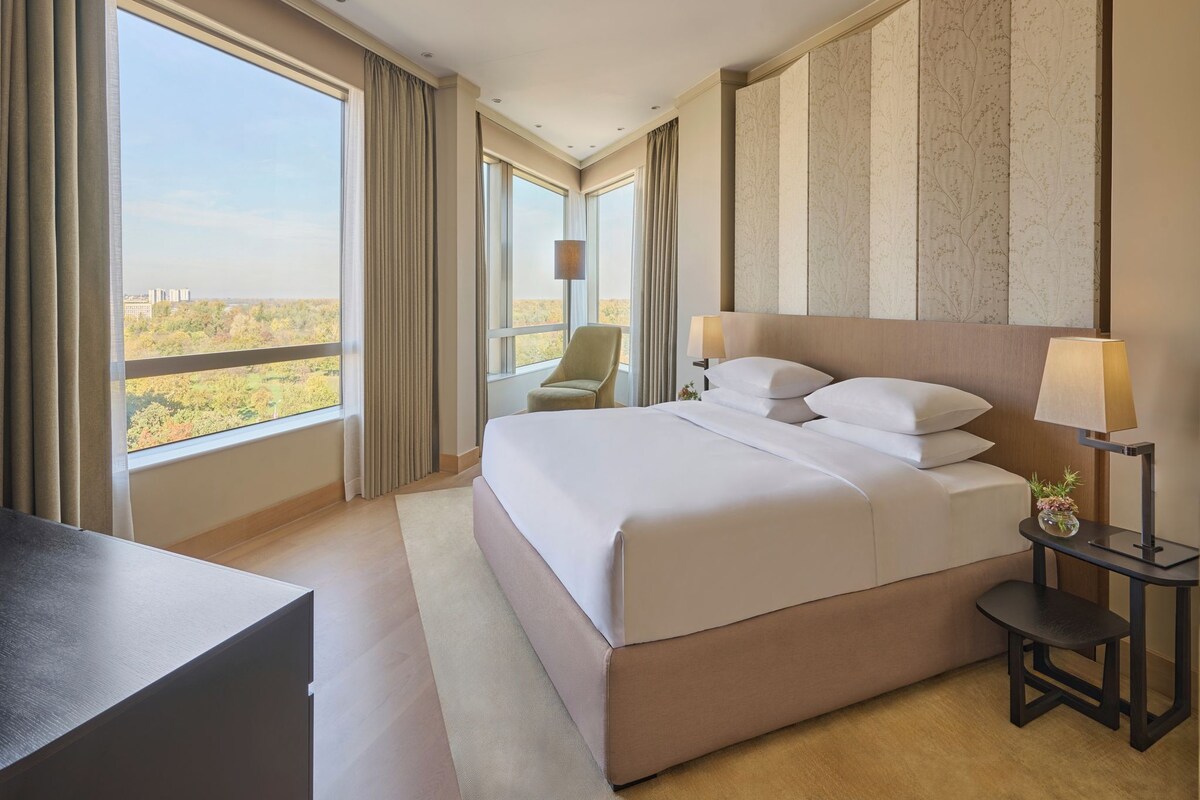 A hotel room with a large bed, a desk, a chair, a nightstand with a lamp, and a view of the cityscape.