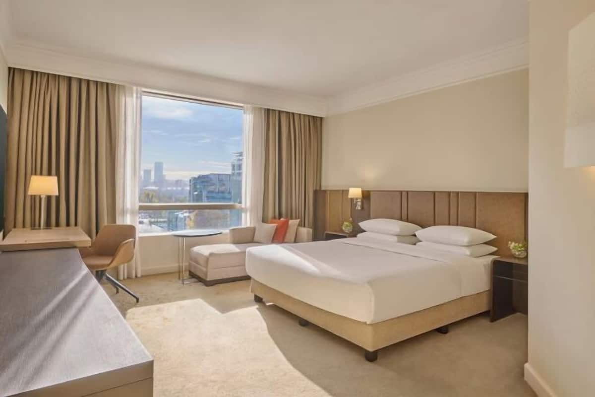A hotel room with a bed, a desk, a chair, a small sofa, and a window with a city view.