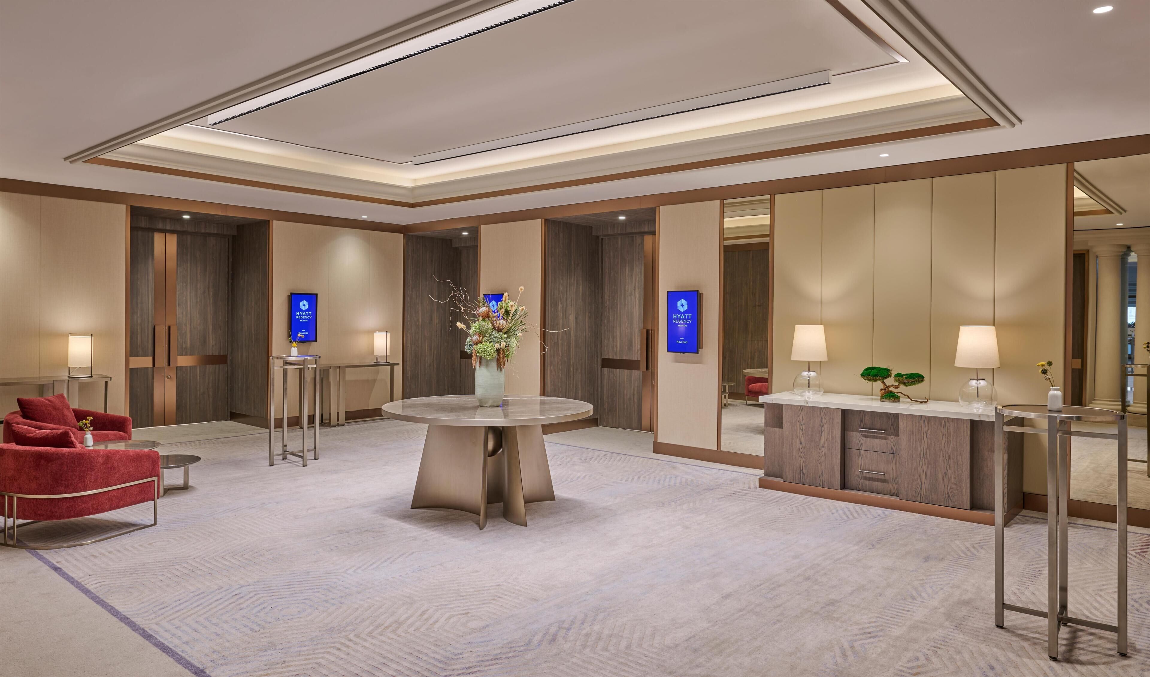 A modern hotel lobby with a round table, red chairs, a reception desk, and a large mirror reflecting the space.