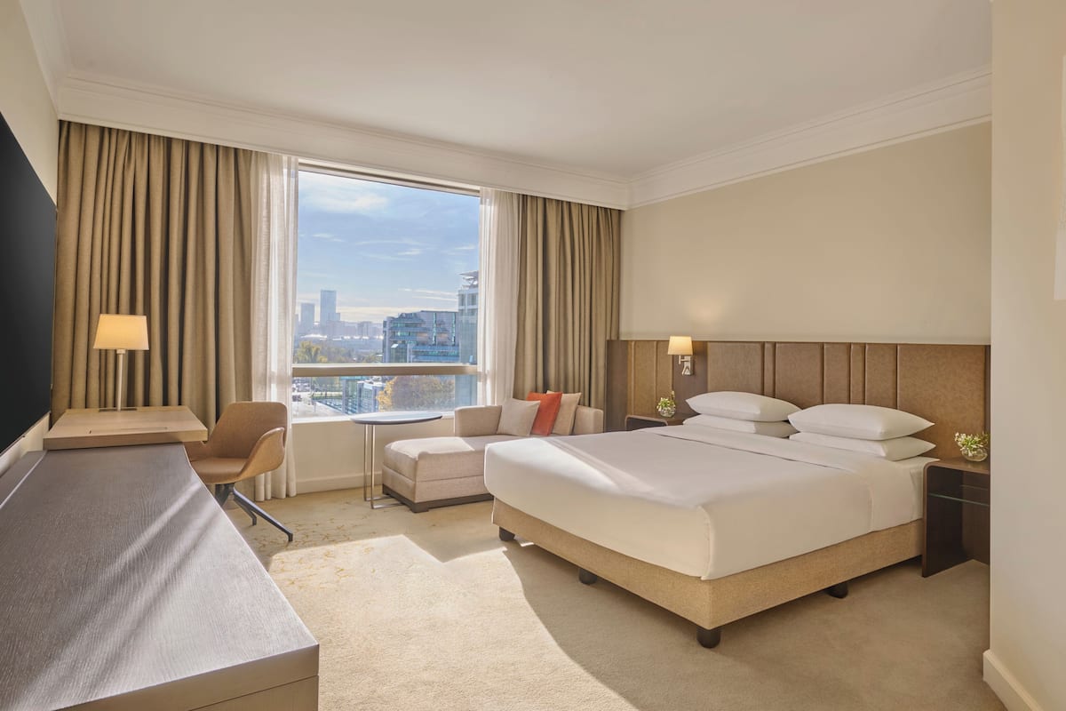 A hotel room with a bed, a desk, a chair, a small table, and a view of the cityscape.