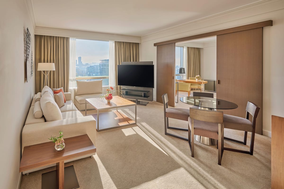 A modern hotel room with a sofa, coffee table, TV, dining area, and city view.