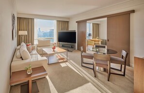 A modern hotel room with a sofa, coffee table, TV, dining area, and city view.