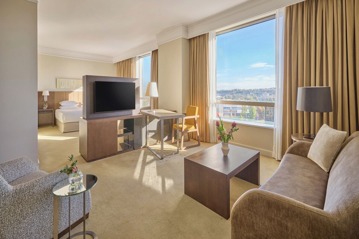 A hotel room with a bed, TV, sofa, armchair, and a table with a vase.
