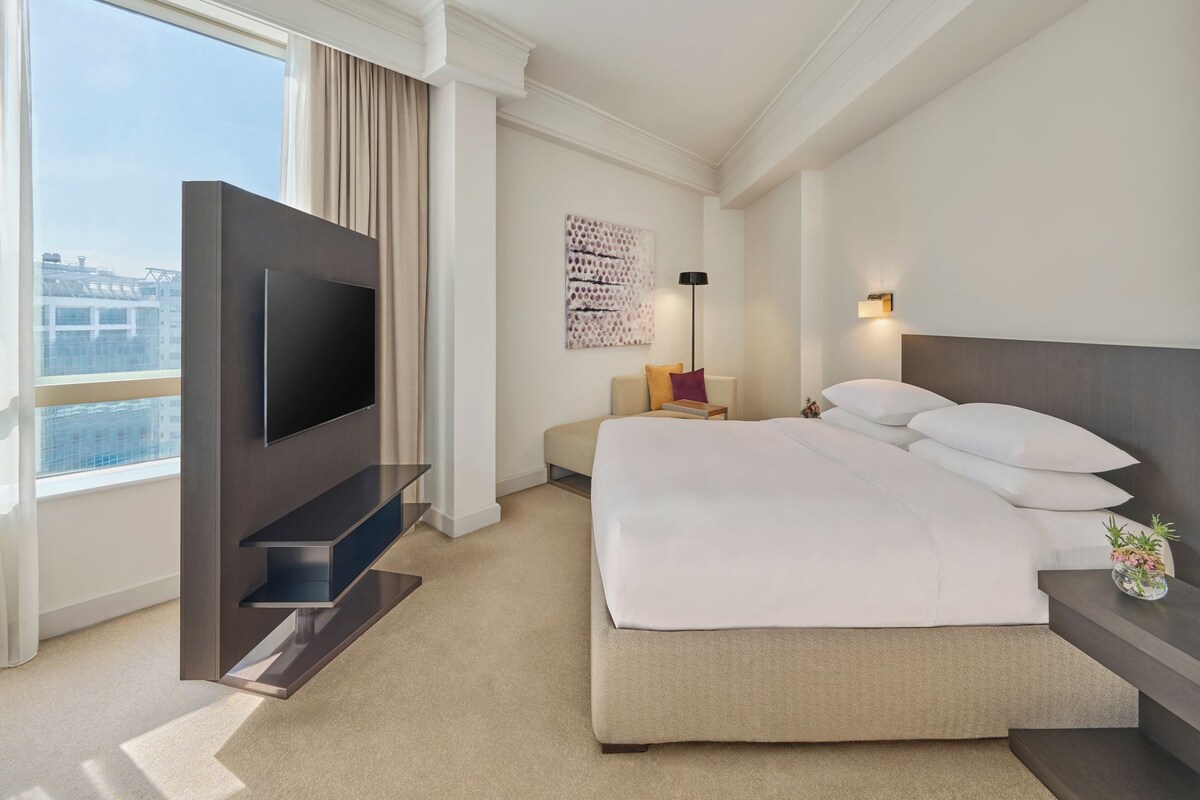 A hotel room with a bed, a TV, a desk, a chair, a lamp, and a view of the city.