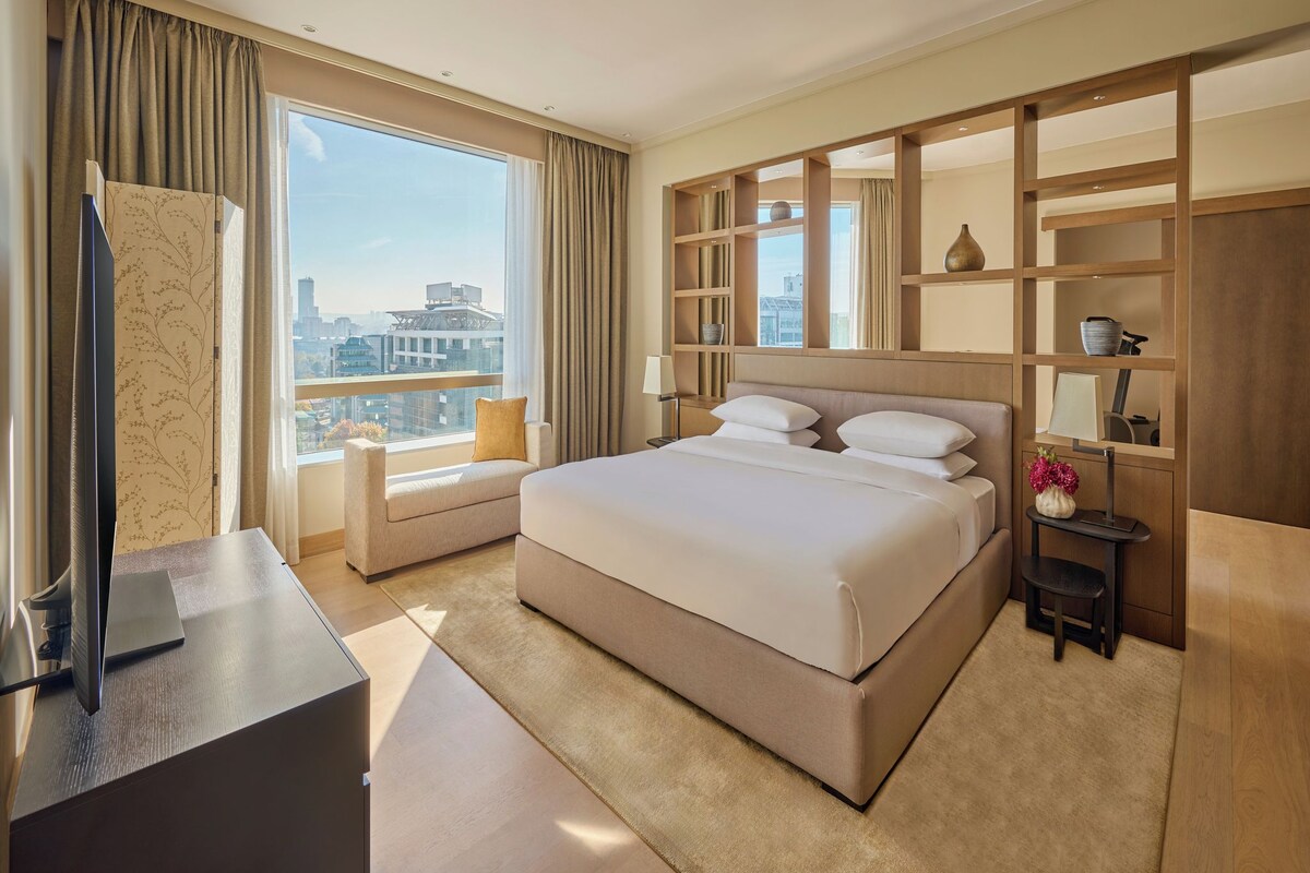 A hotel room with a large bed, a desk with a laptop, a window with curtains, and a city view.