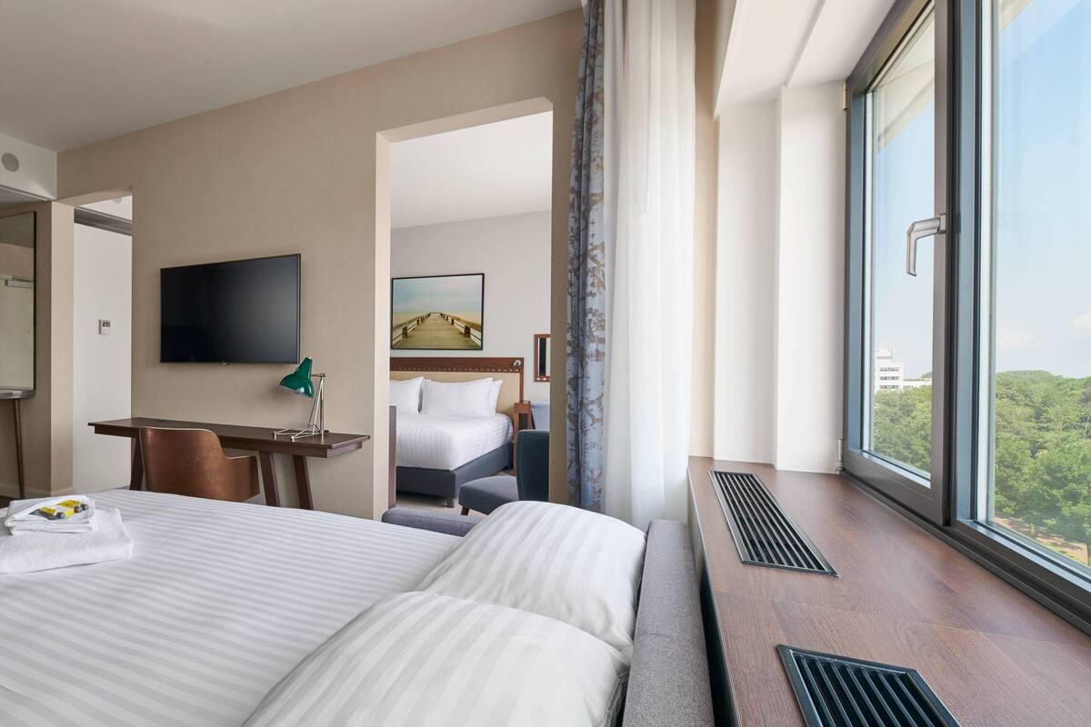 A hotel room with two beds, a desk, a TV, and a large window with a view.