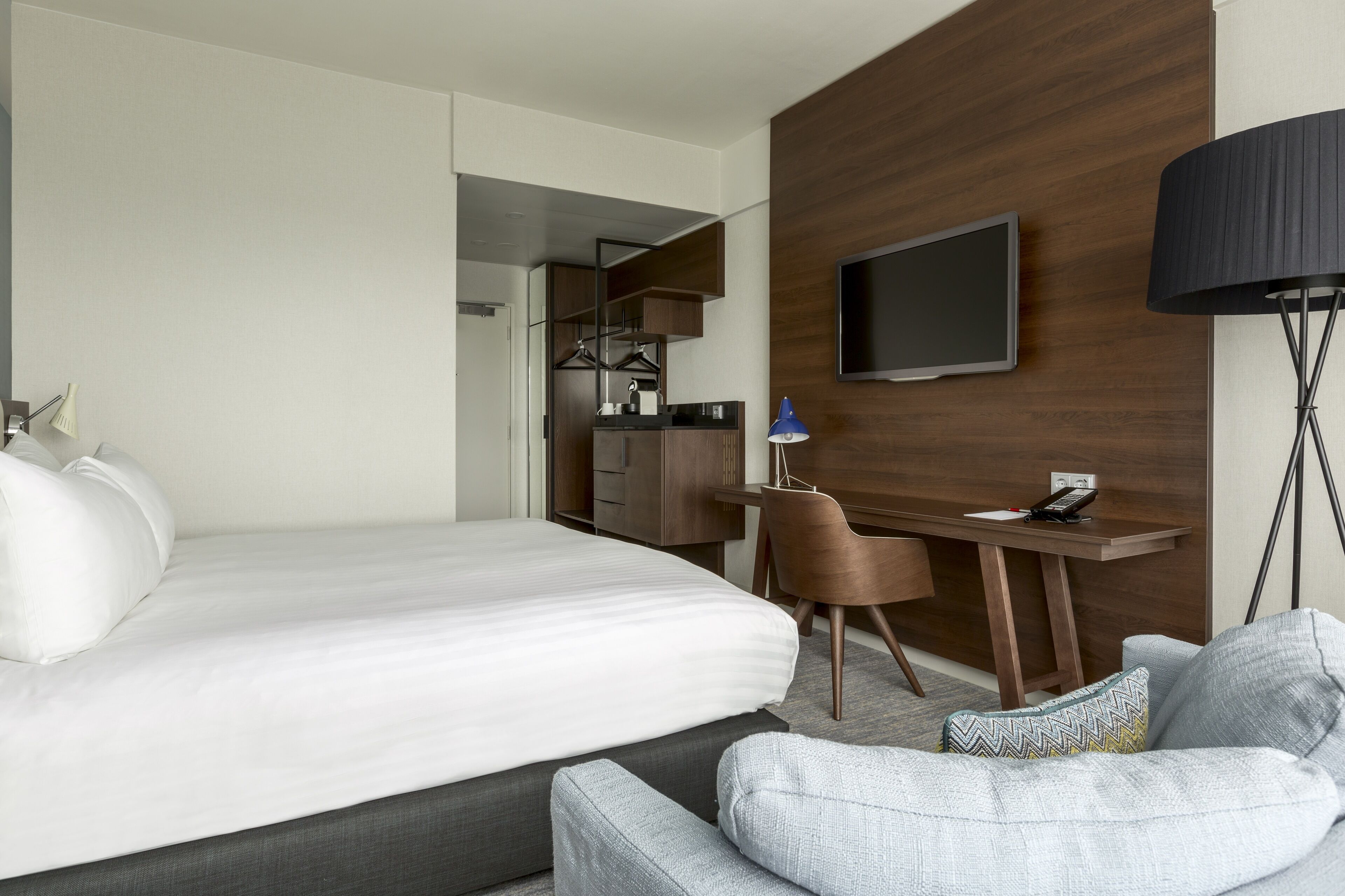 A modern hotel room with a large bed, a desk, a chair, a TV, and a lamp.