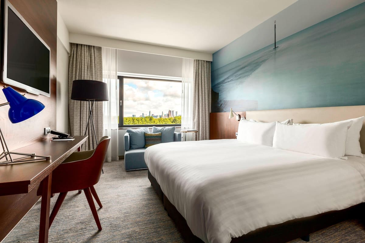 A hotel room with a large bed, a desk with a chair, a sofa, and a city view through the window.