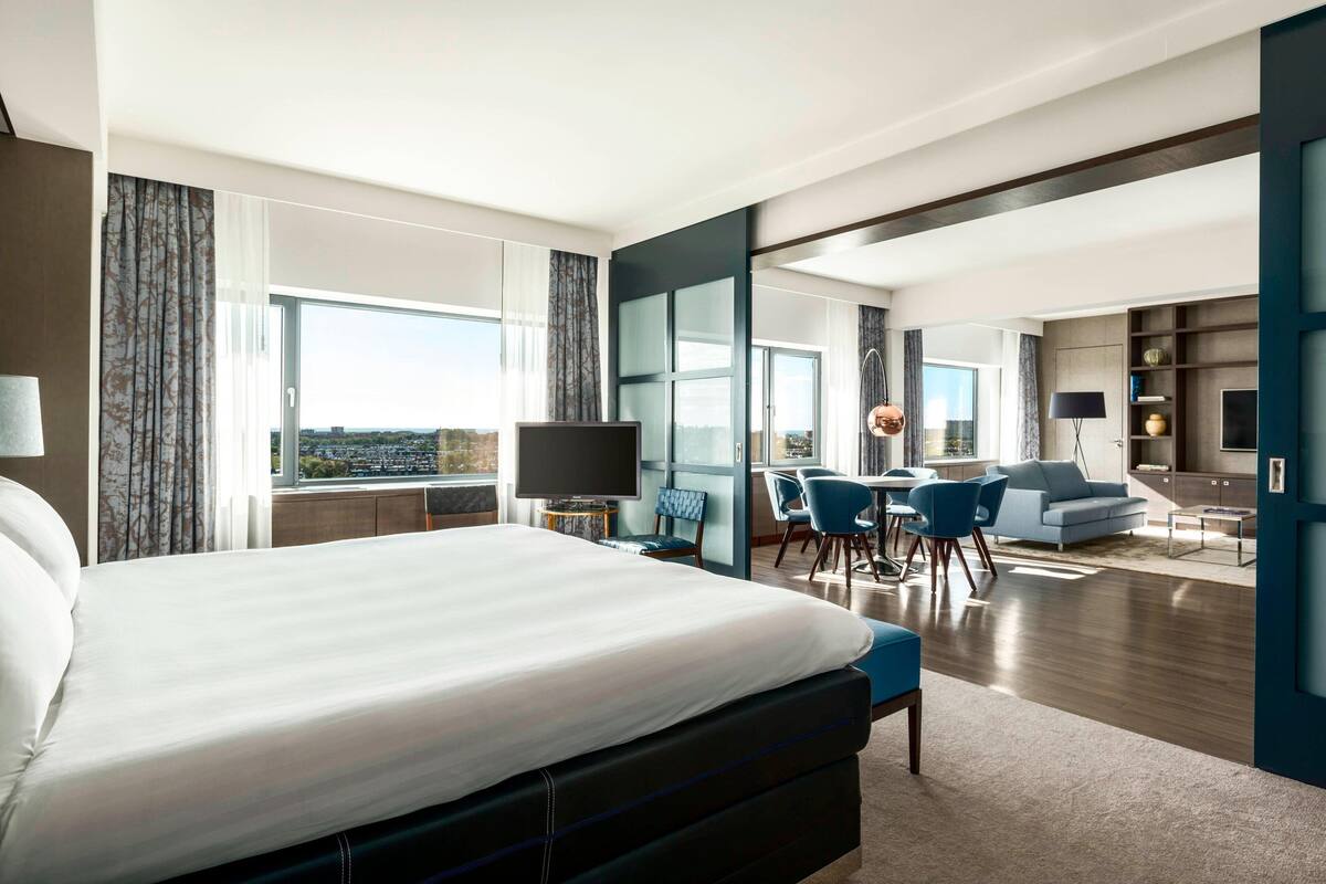 A modern hotel room with a large bed, a TV, a dining area with chairs, and a sofa.