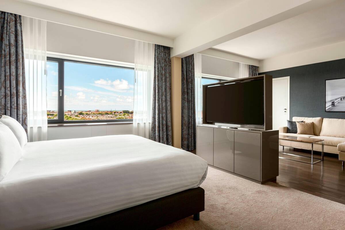 A modern hotel room with a large bed, a TV, a sofa, and a view of the city.