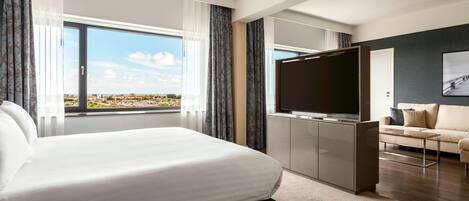A modern hotel room with a large bed, a TV, a sofa, and a view of the city.
