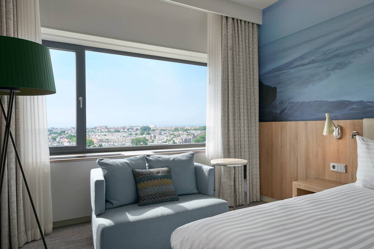 A hotel room with a large window offering a city view, a sofa with a patterned pillow, a bedside table, and a wall-mounted lamp.