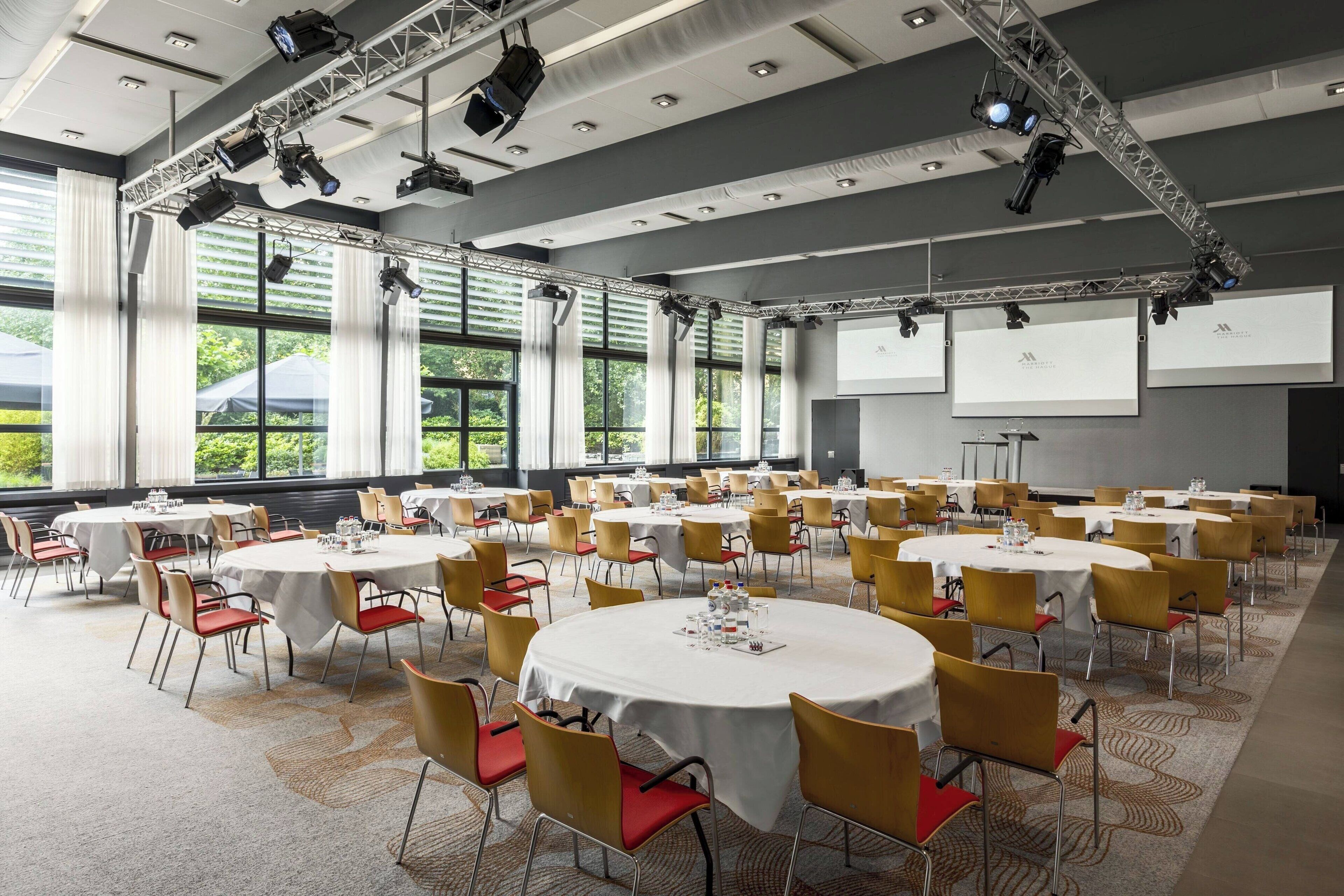 A conference room with multiple round tables, chairs, and a stage with lighting equipment.