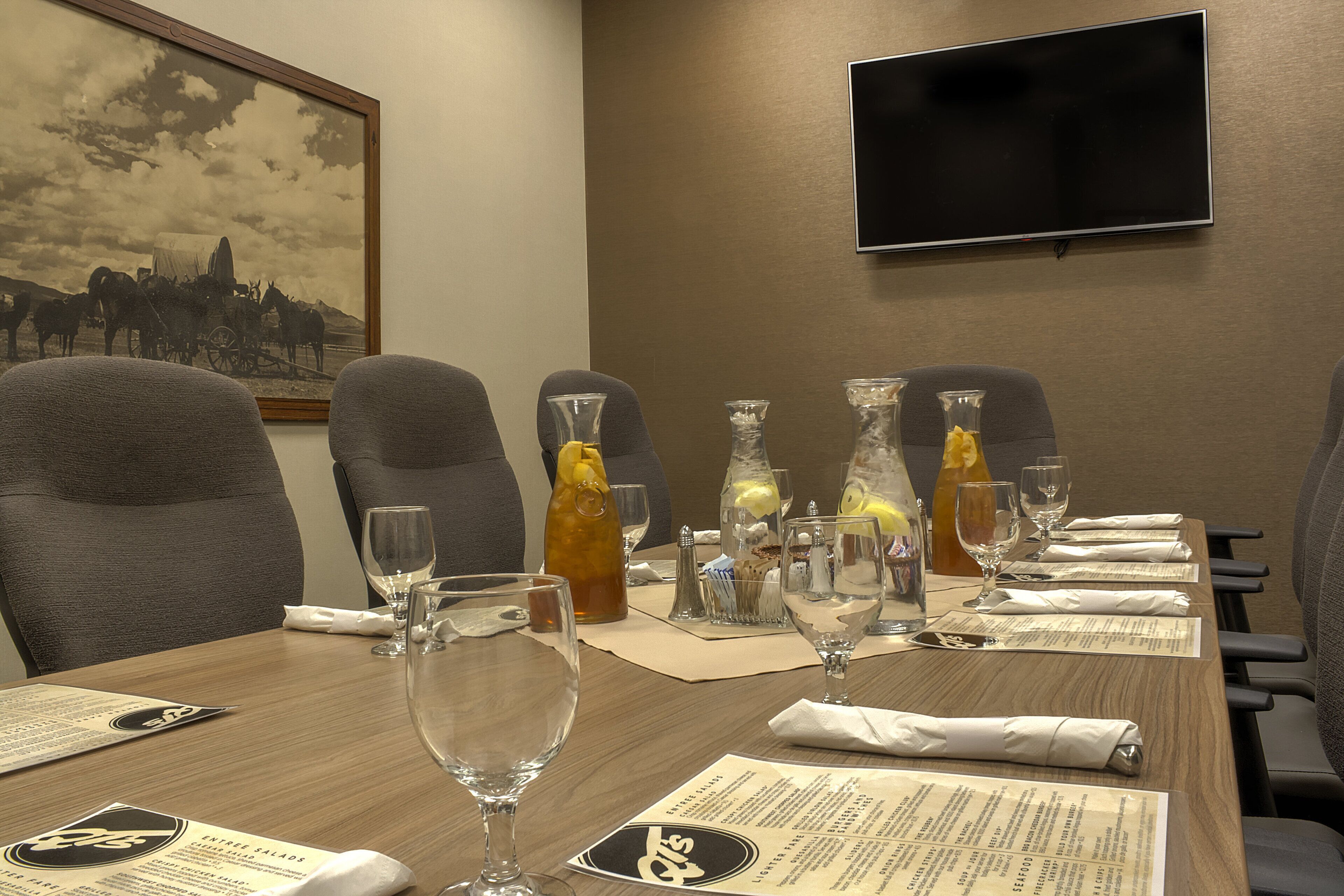 Een vergaderzaal met een houten tafel gedekt voor een vergadering, een aan de muur bevestigde televisie en een in een lijst gezet historisch schilderij.