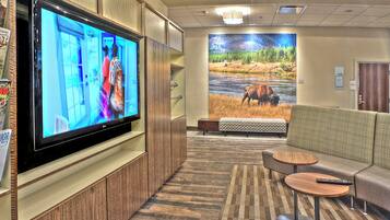 A hotel lobby with a large flat-screen TV mounted on a wooden cabinet, a seating area with a sofa and two small round tables, and a large mural of a river scene on the back wall.