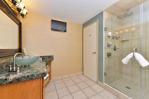 Un baño con ducha de vidrio, un lavabo con encimera de mármol y un televisor de pantalla plana montado en la pared.