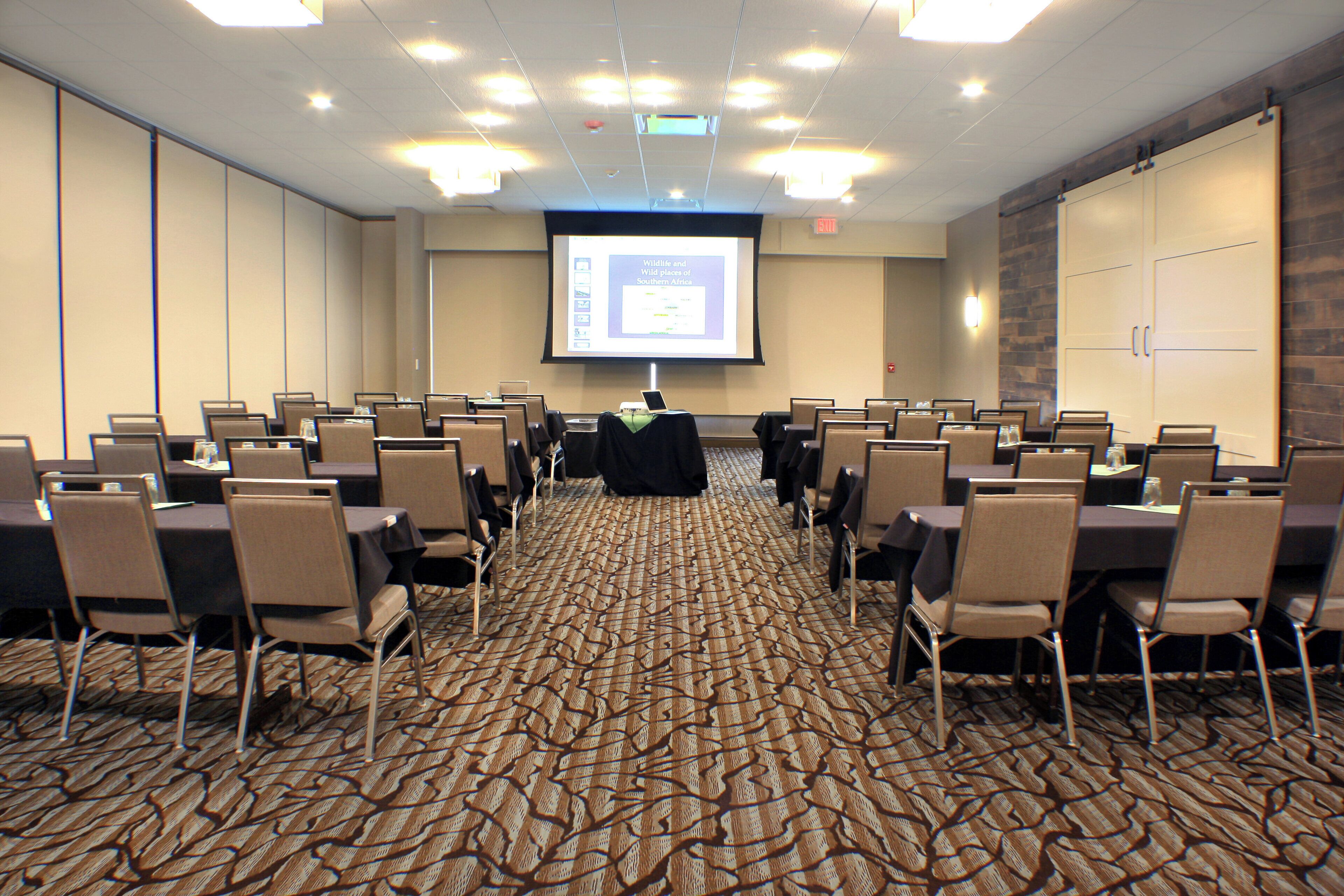 A conference room with rows of chairs and tables, a projector screen displaying a presentation, and a carpeted floor.
