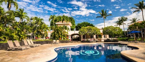 Una piscina con sillas de descanso, palmeras y un cielo azul despejado.