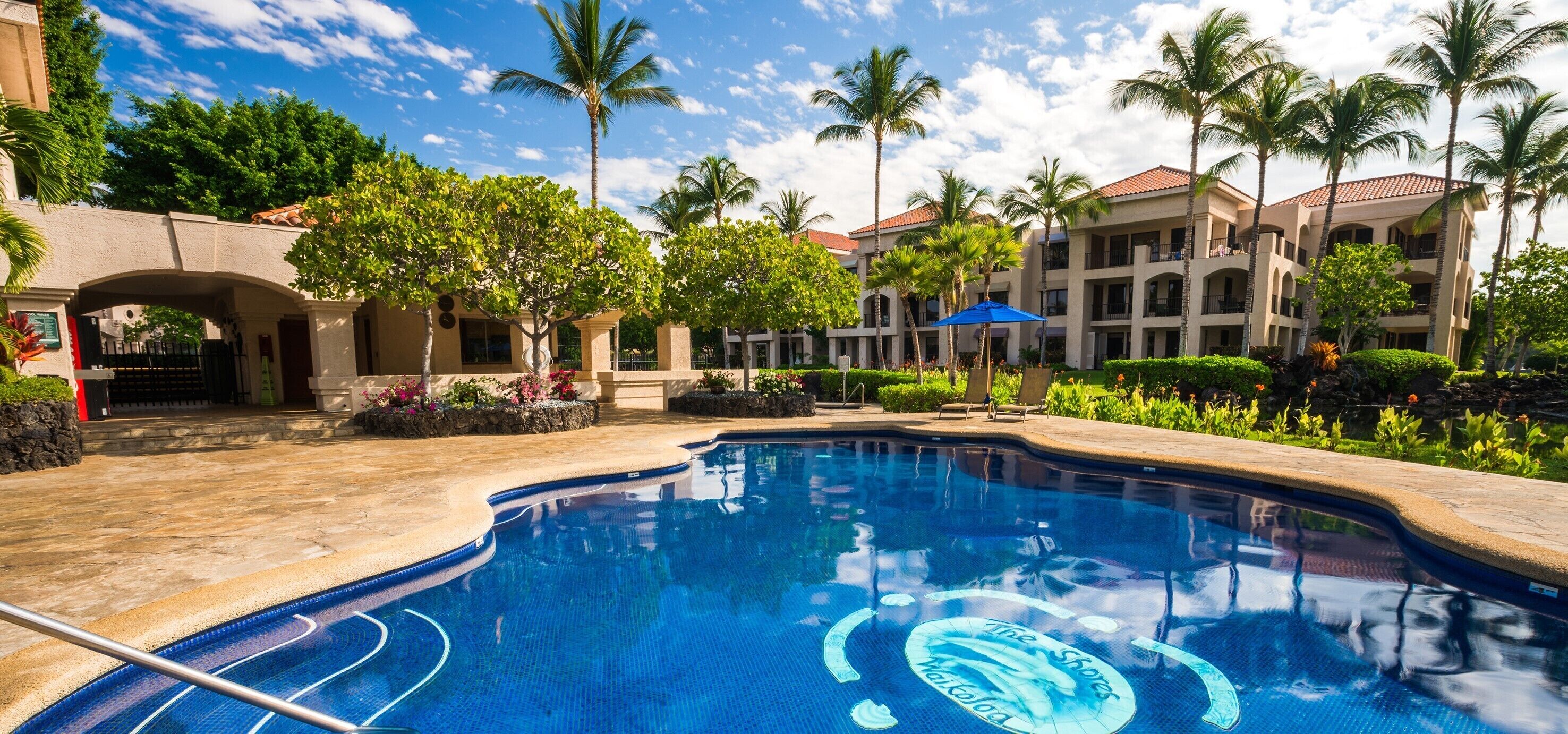 A swimming pool with a clear blue water feature, surrounded by palm trees and a multi-story building with balconies.
