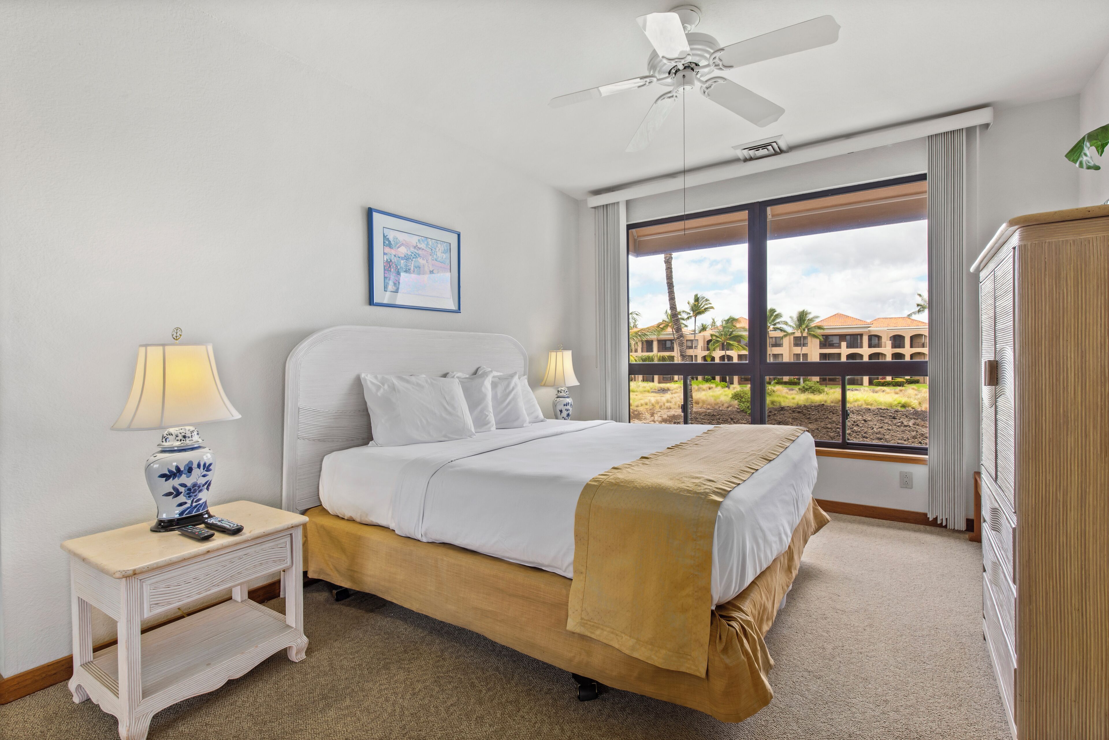 A neatly arranged hotel room with a bed, bedside lamps, a ceiling fan, and a window offering a view of palm trees and buildings.