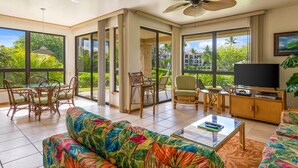 A spacious living room with large windows, a ceiling fan, a television, and a sofa with a tropical print. - Shores at Waikoloa (Waikoloa)