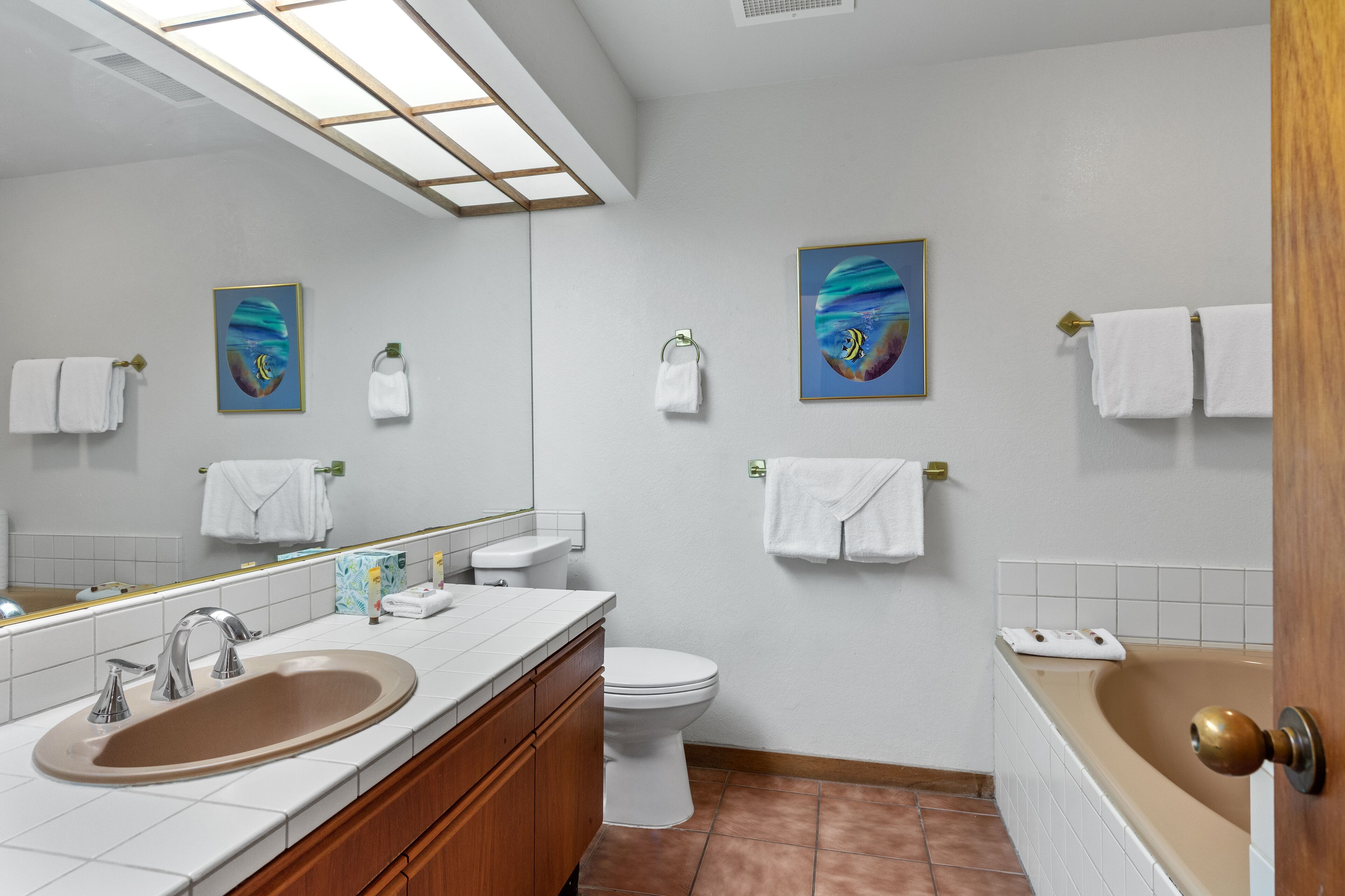 Un baño moderno con lavamanos, inodoro y bañera. Dos cuadros enmarcados adornan las paredes, y varias toallas blancas cuelgan de barras.
