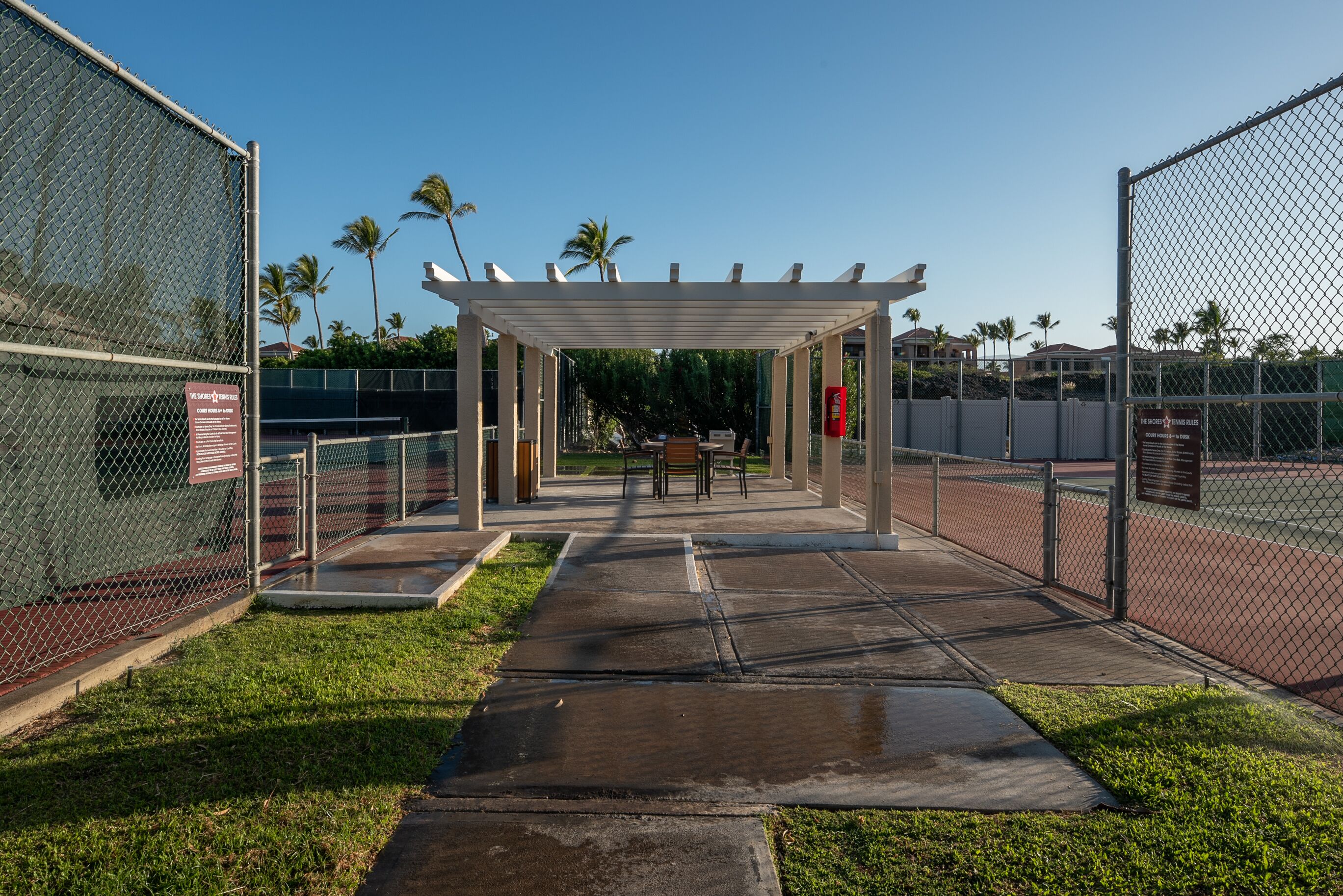 A tennis court with a covered seating area, surrounded by chain-link fences and palm trees.