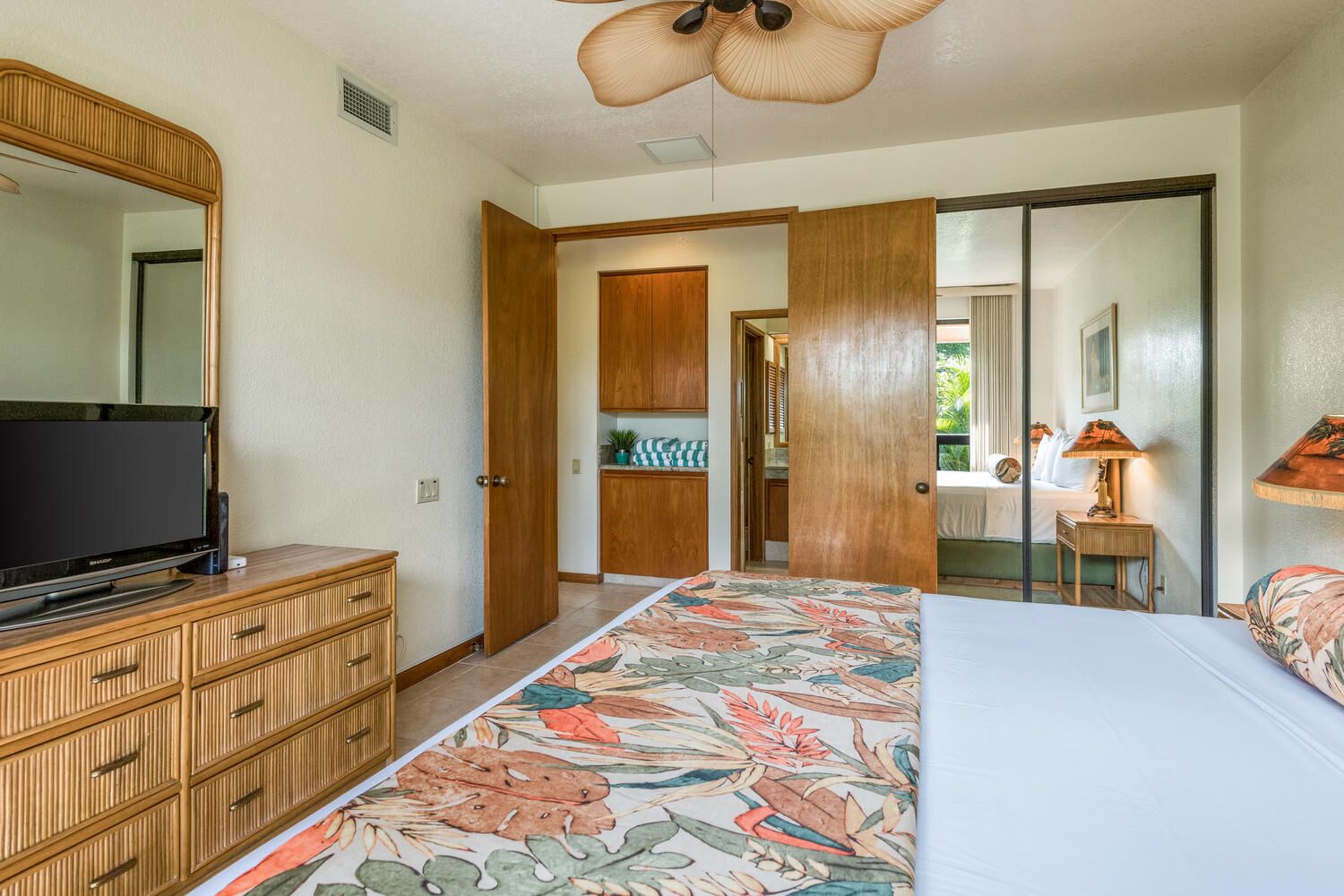 Une chambre à coucher avec un lit, un téléviseur posé sur une commode en bois, une porte donnant sur une salle de bain et un ventilateur de plafond.