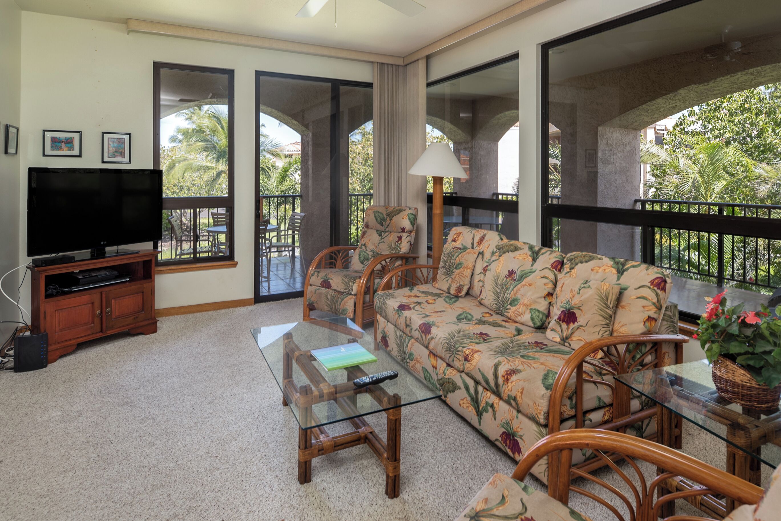 A living room with a sofa, a coffee table, a TV, and a balcony with a view of palm trees.