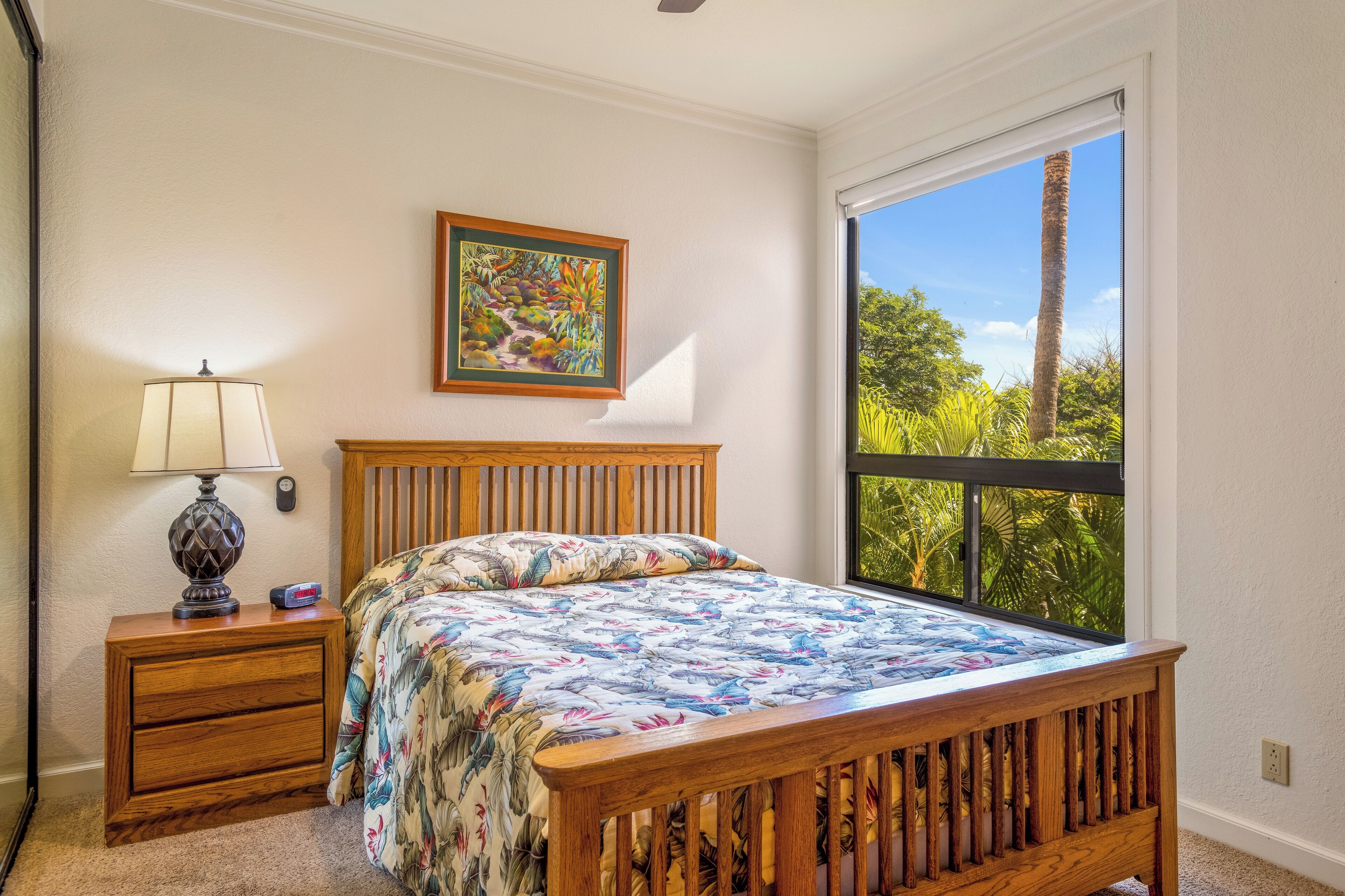 A bedroom with a wooden bed, a nightstand with a lamp, a framed picture on the wall, and a large window with a view of greenery.