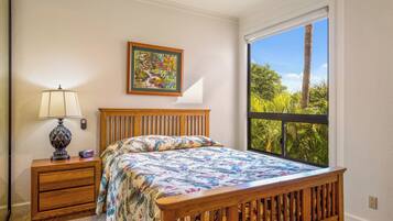 A bedroom with a wooden bed, a nightstand with a lamp, a framed picture on the wall, and a large window with a view of greenery.