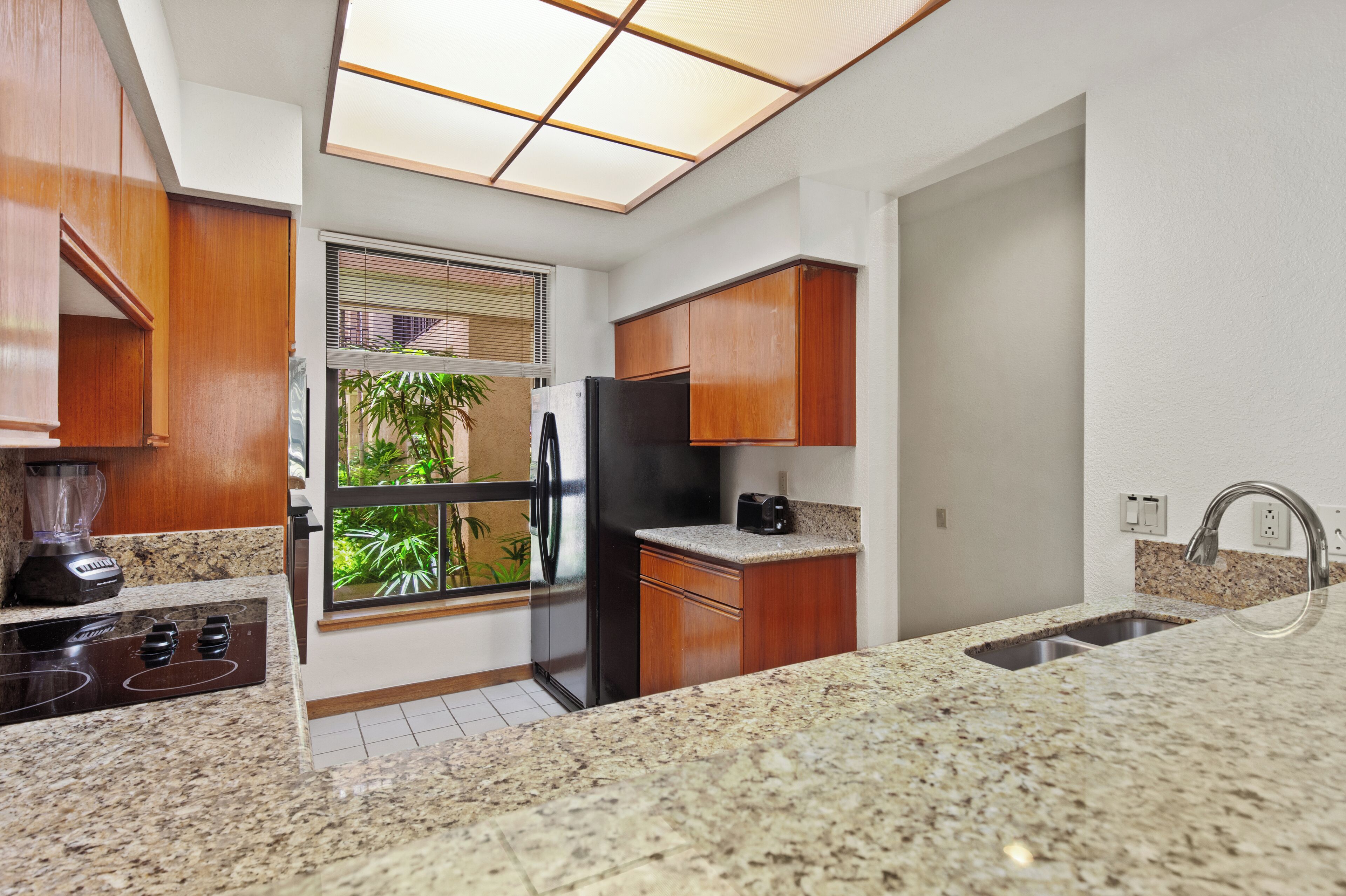 A kitchen with granite countertops, wooden cabinets, a stainless steel refrigerator, and a window with blinds.