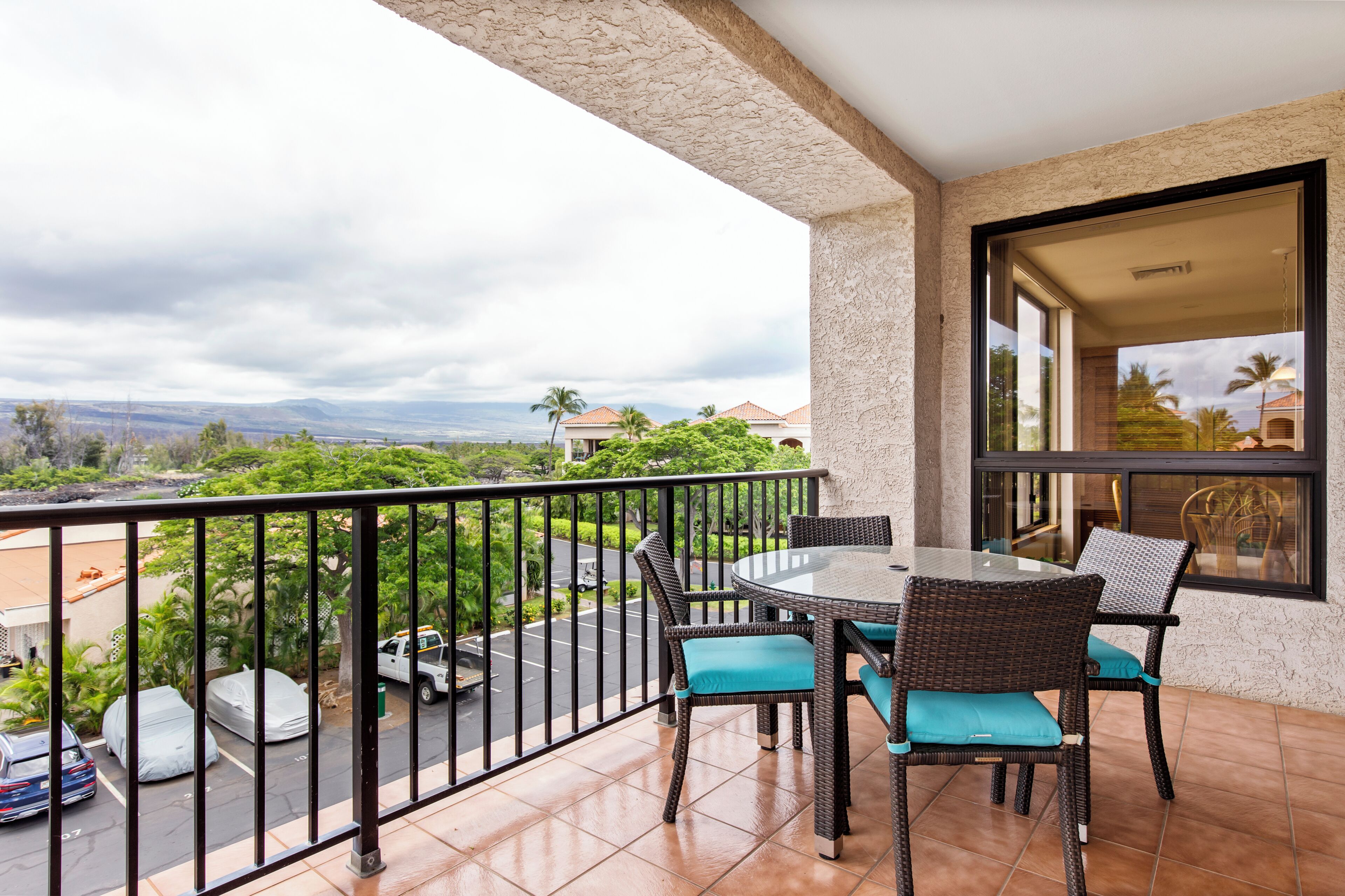 A balcony with a table and chairs, a view of a parking lot, and a building with a window.