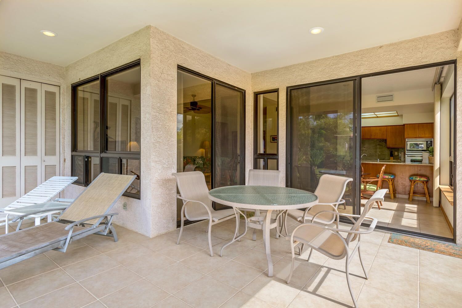 A sunlit patio with a round table and chairs, a lounge chair, and a sliding glass door leading to a kitchen area.