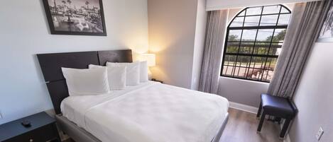 Una cama bien hecha con ropa blanca, una mesita de noche con un control remoto, una ventana con cortinas y un cuadro en la pared.