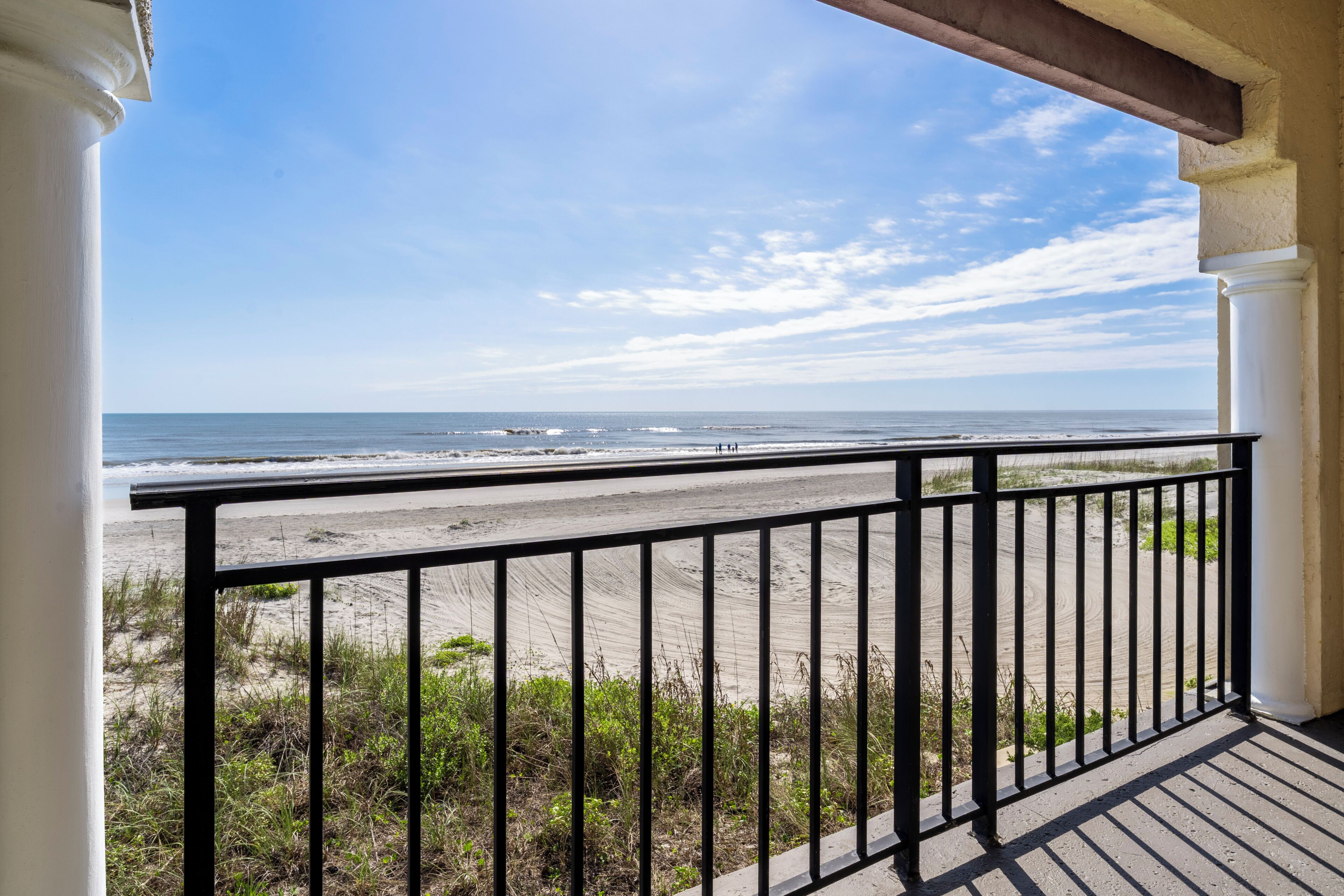 A balcony with a view of the beach, white columns, and a clear sky.