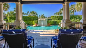 A covered outdoor area with a pool, lounge chairs, and palm trees.