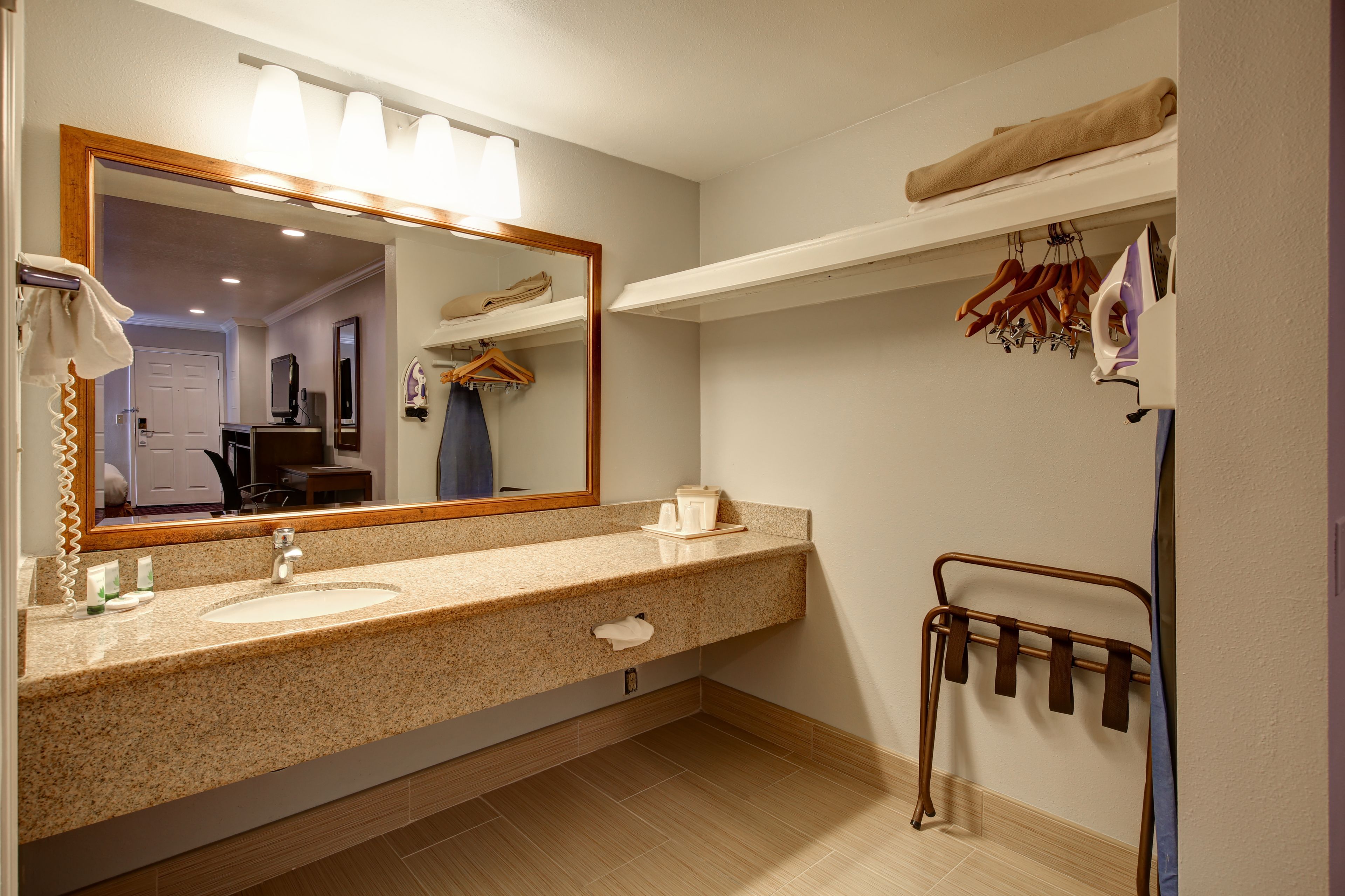 A bathroom with a granite countertop sink, a large mirror with lights, a towel rack, and a shelf with towels and hangers.