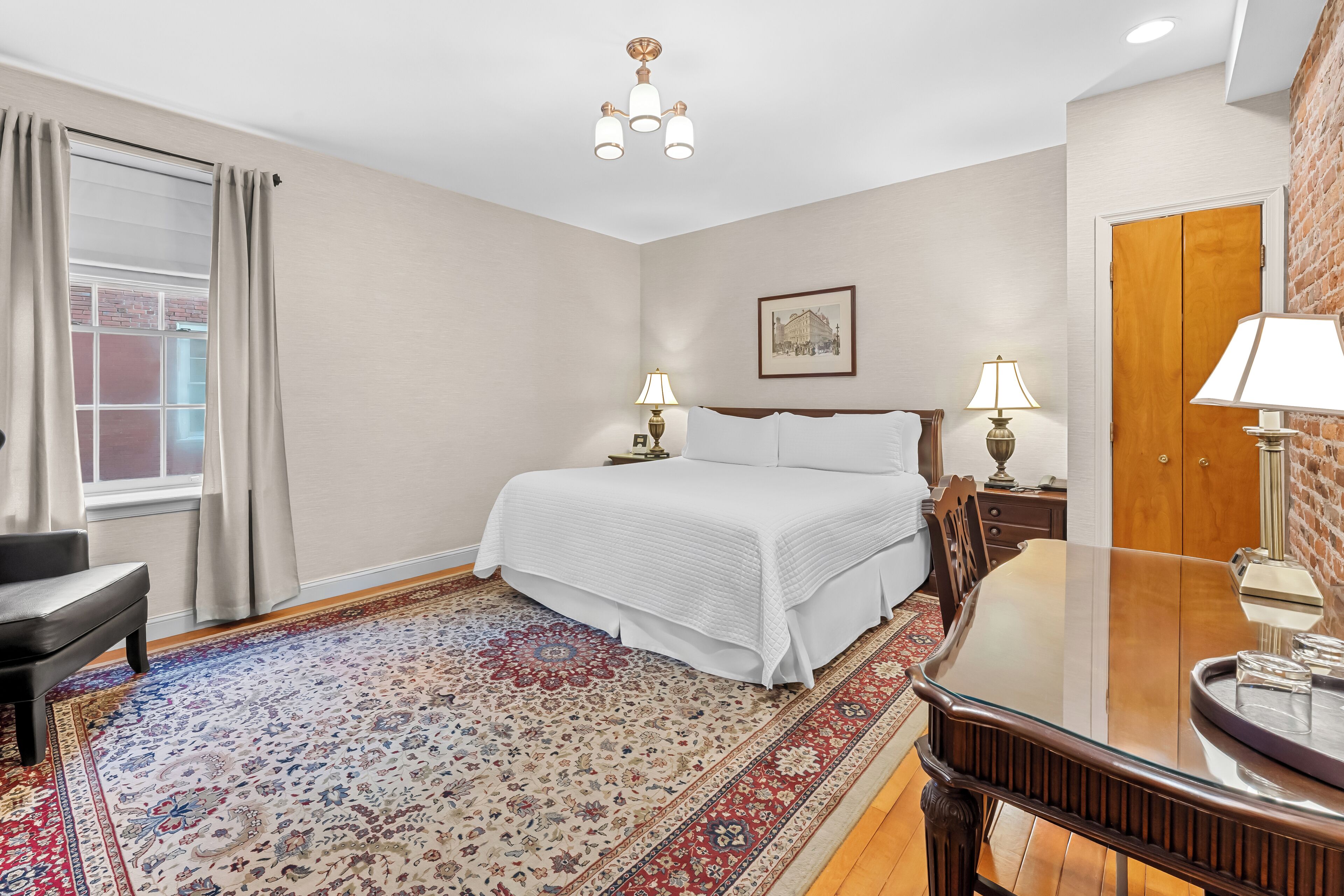 Een hotelkamer met een groot bed, twee bedlampen, een houten vloer, een kamerbreed tapijt met een patroon, een raam met gordijnen en een deur met spiegel.