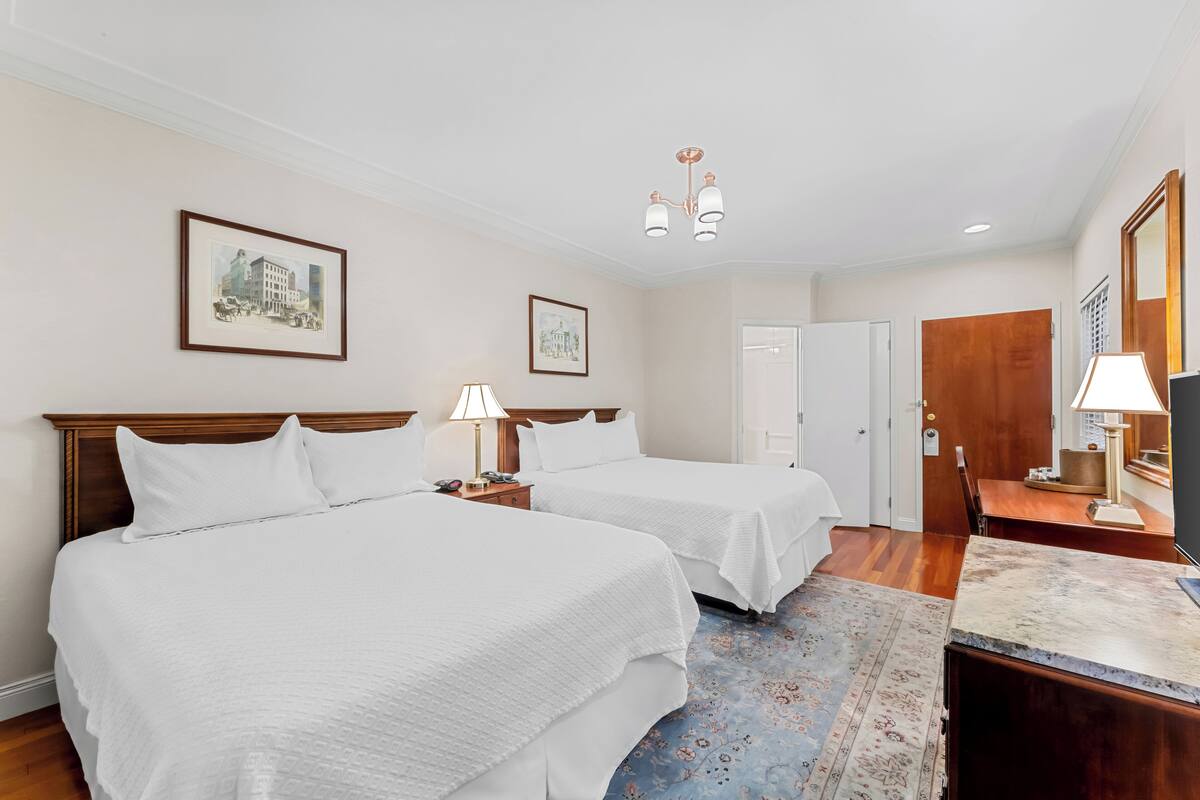 A hotel room with two beds, a desk, a chair, a lamp, a mirror, and two framed pictures on the wall.
